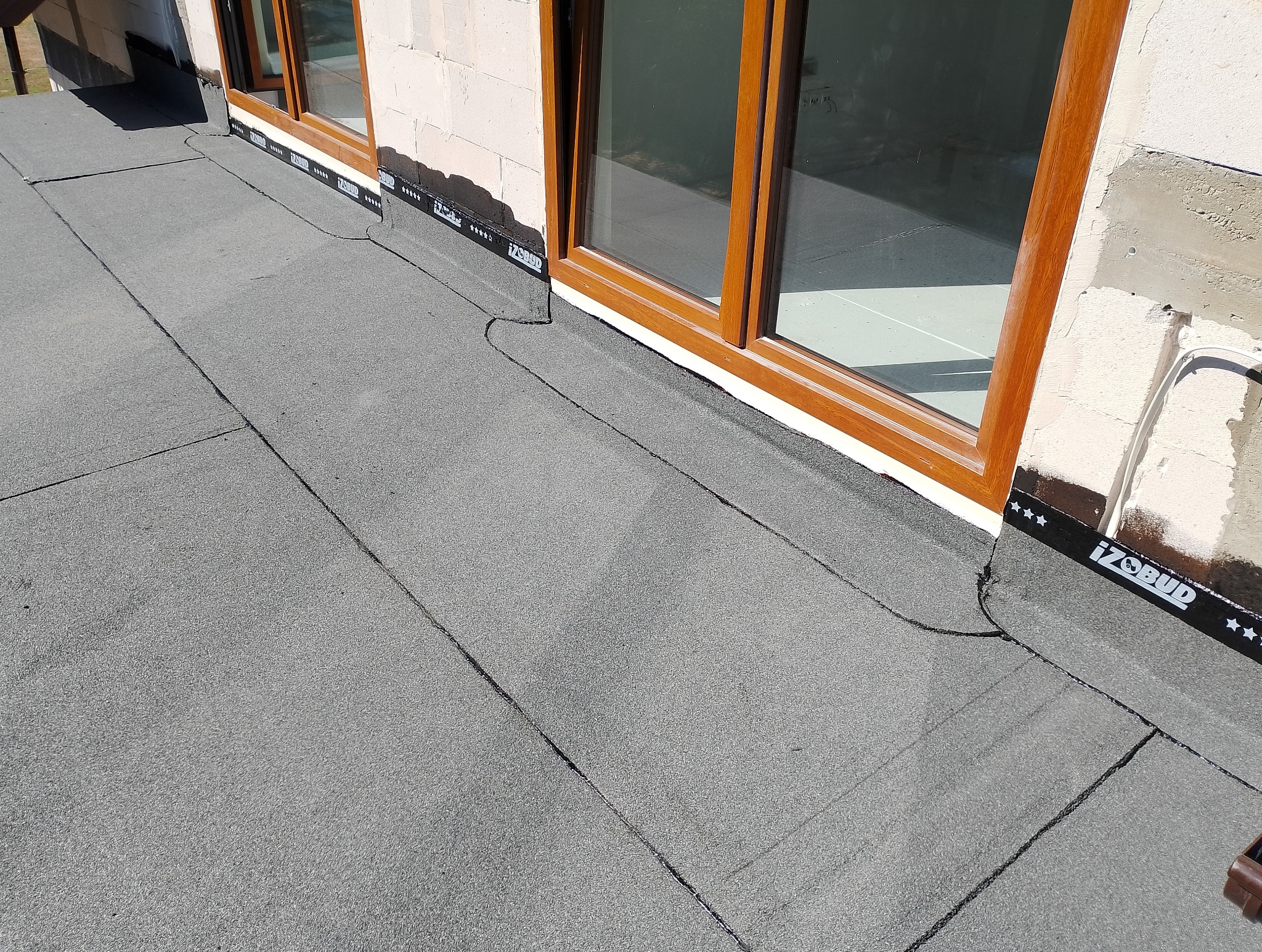 Płaski dach pokryty papą termozgrzewalną, z widocznymi oknami dachowymi i fragmentem ściany budynku z elementami wykończeniowymi, uszczelniony taśmą marki Izobud.