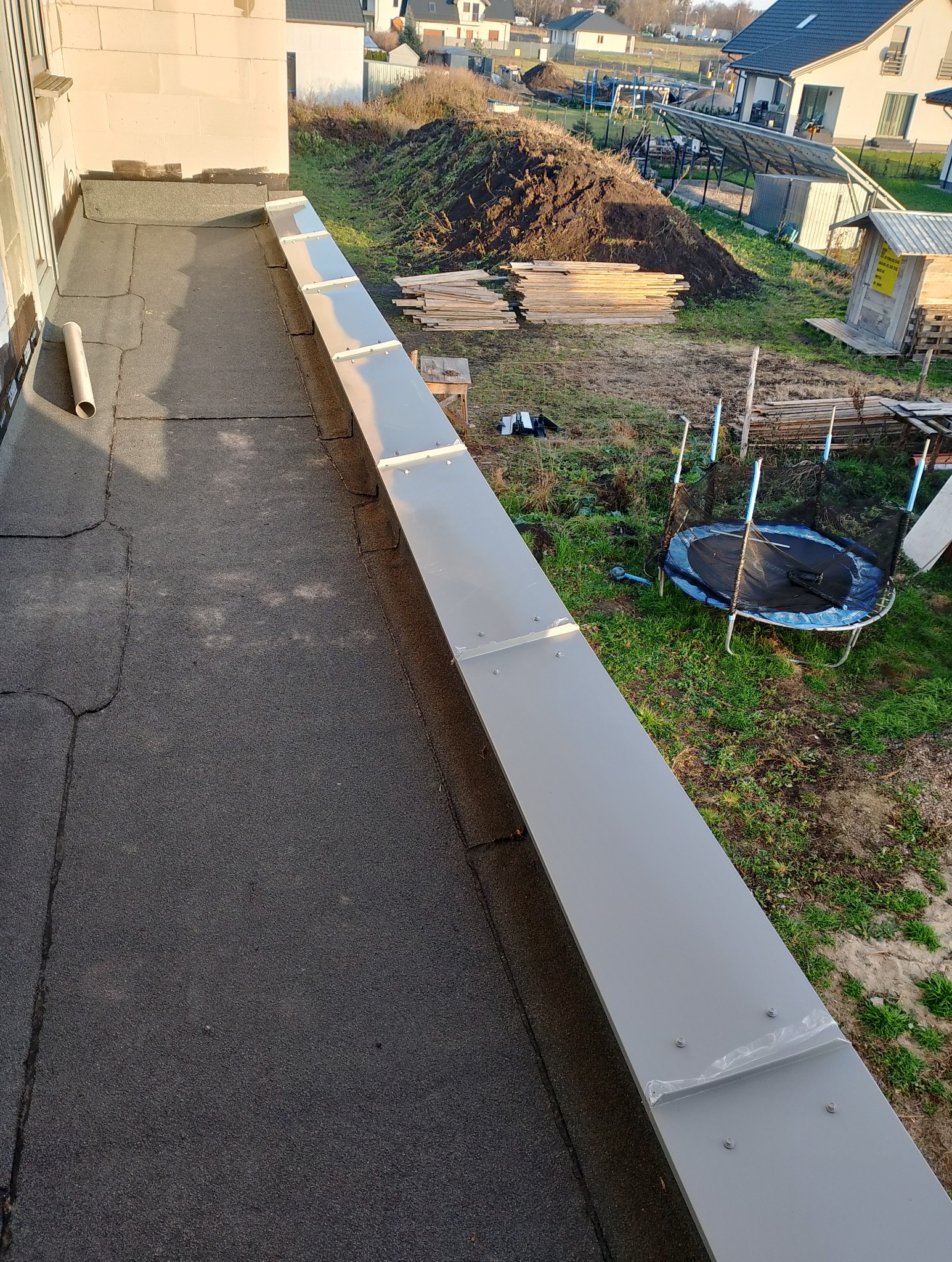 Widok z góry na fragment dachu pokrytego papą z nowo zamontowaną, długą, szarą obróbką blacharską, zabezpieczającą krawędź dachu; w tle trawnik z trampoliną i stertą desek.