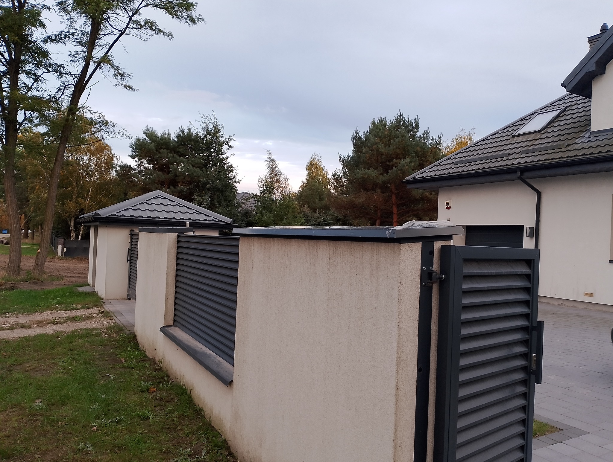 Nowoczesne ogrodzenie posesji z metalowymi przęsłami w kolorze grafitowym i jasnymi, otynkowanymi słupkami, widok na dom z grafitowym dachem i rynnami.