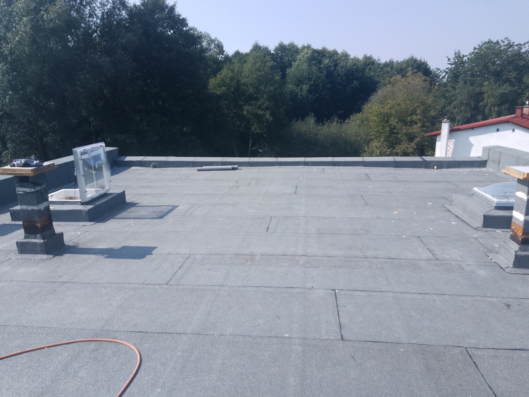 Szare, płaskie pokrycie dachowe z papy termozgrzewalnej na dachu budynku, widoczne kominki wentylacyjne i świetliki dachowe, w tle drzewa i fragment budynku z czerwonym dachem.