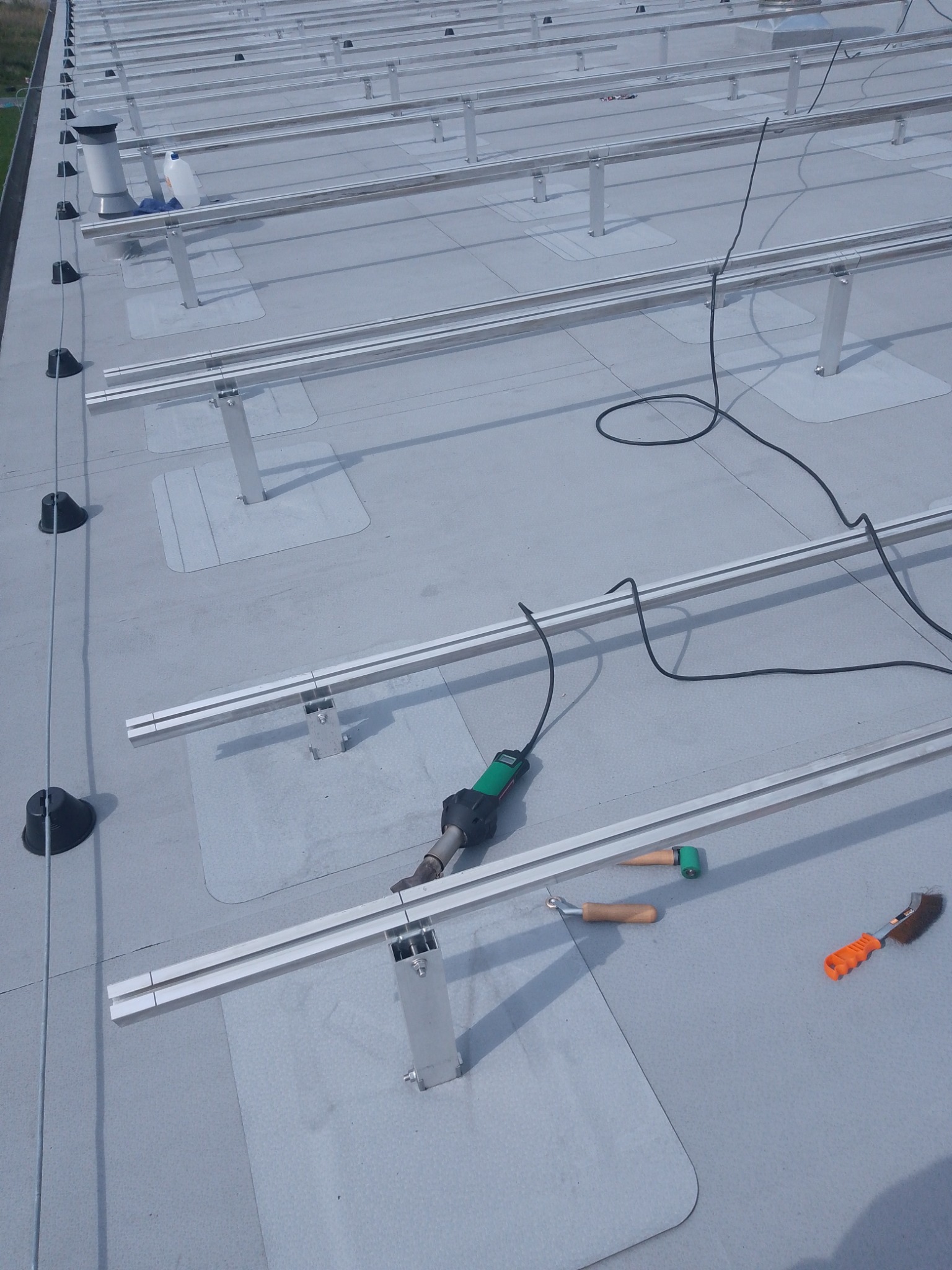 Montaż aluminiowej konstrukcji wsporczej pod panele fotowoltaiczne na dachu pokrytym membraną, widoczne narzędzia: zgrzewarka do membran, dociskacz, szczotka.
