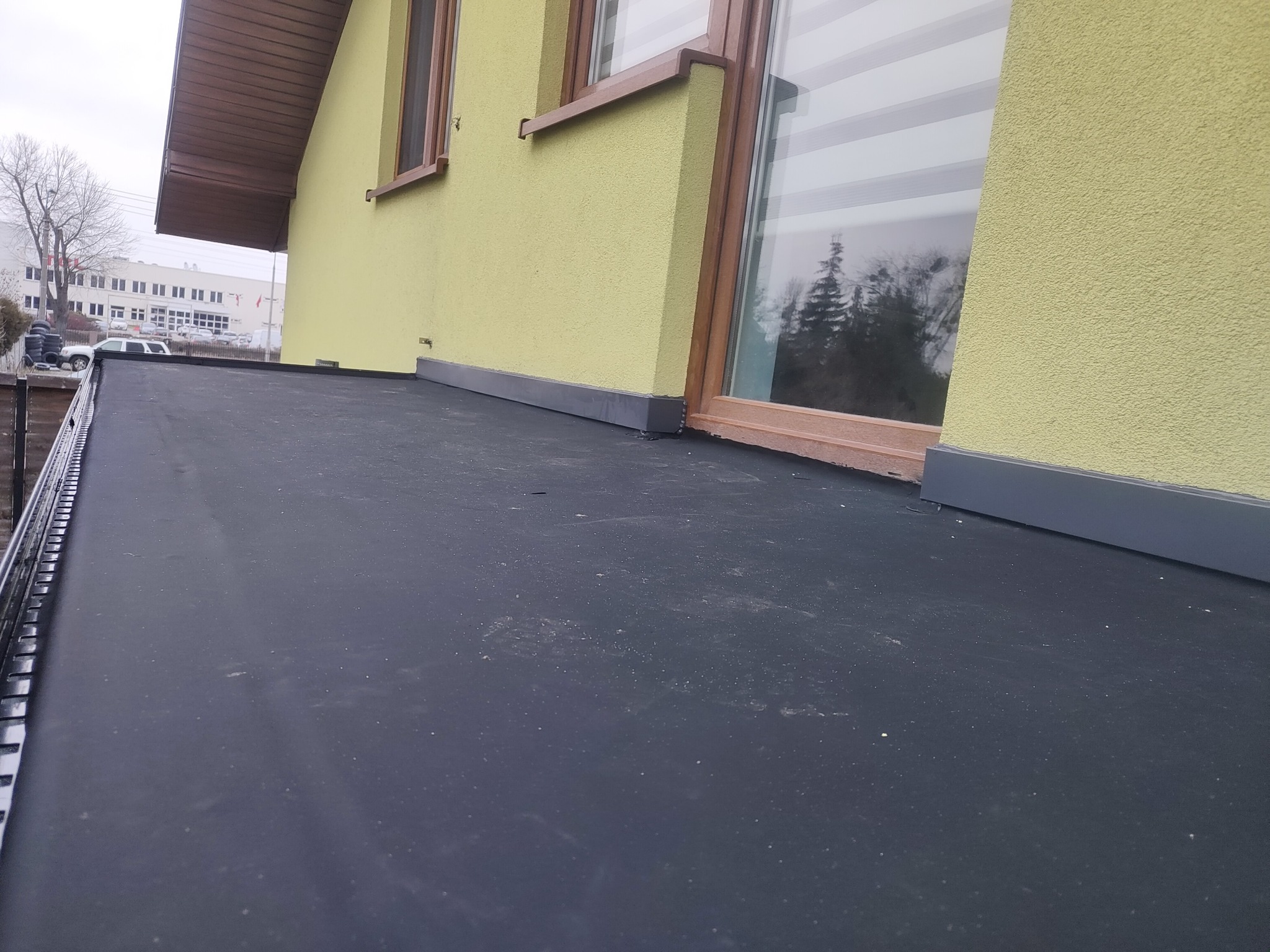 Widok z bliska na płaski dach pokryty czarną membraną bitumiczną, z widoczną obróbką blacharską przy ścianie budynku o żółtej elewacji i oknach z brązowymi ramami. W tle budynek przemysłowy...