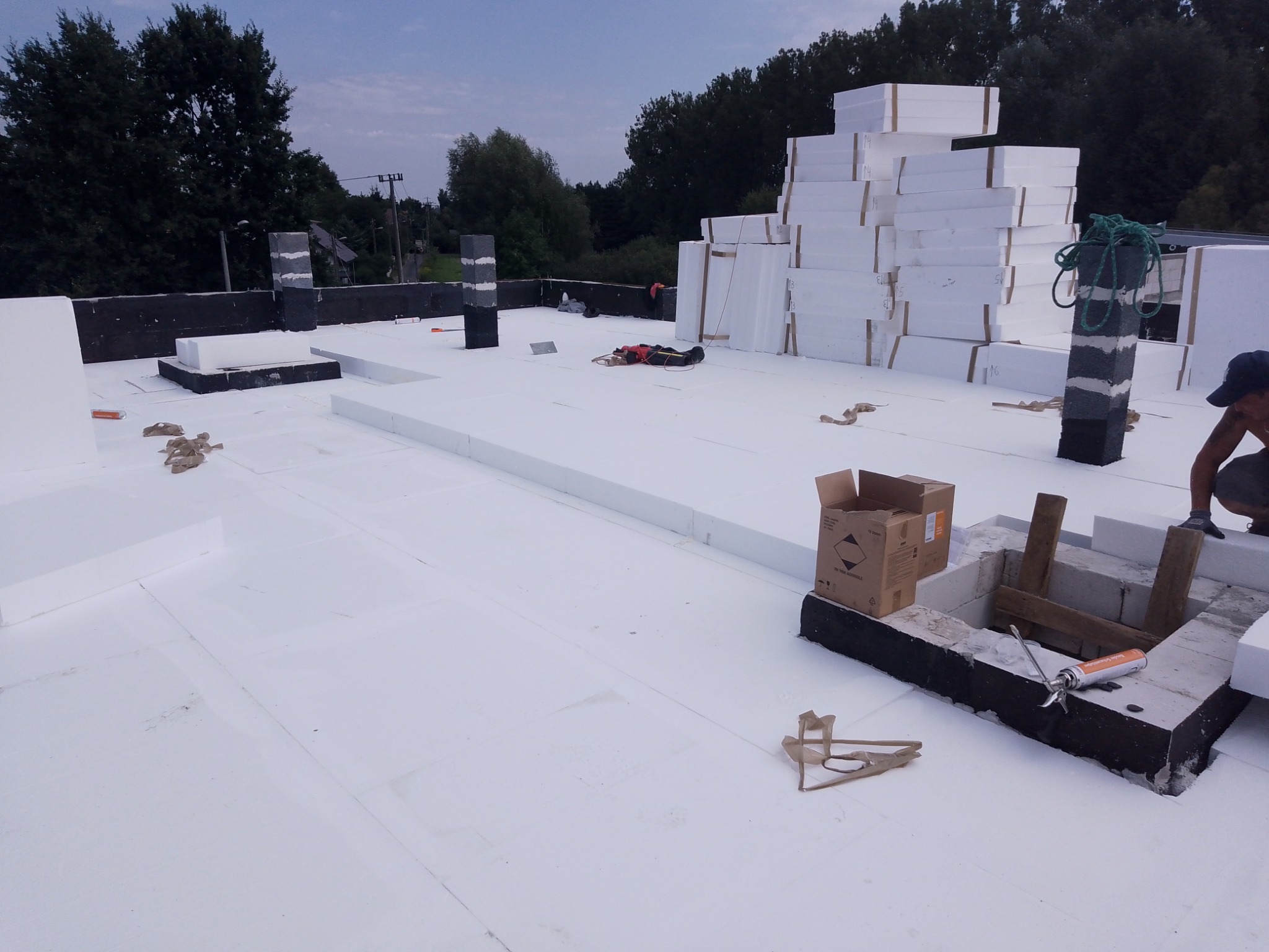 Białe płyty styropianowe ułożone na dachu, widoczny pracownik w trakcie pracy, narzędzia i materiały budowlane rozłożone na powierzchni dachu, w tle drzewa i niebo.