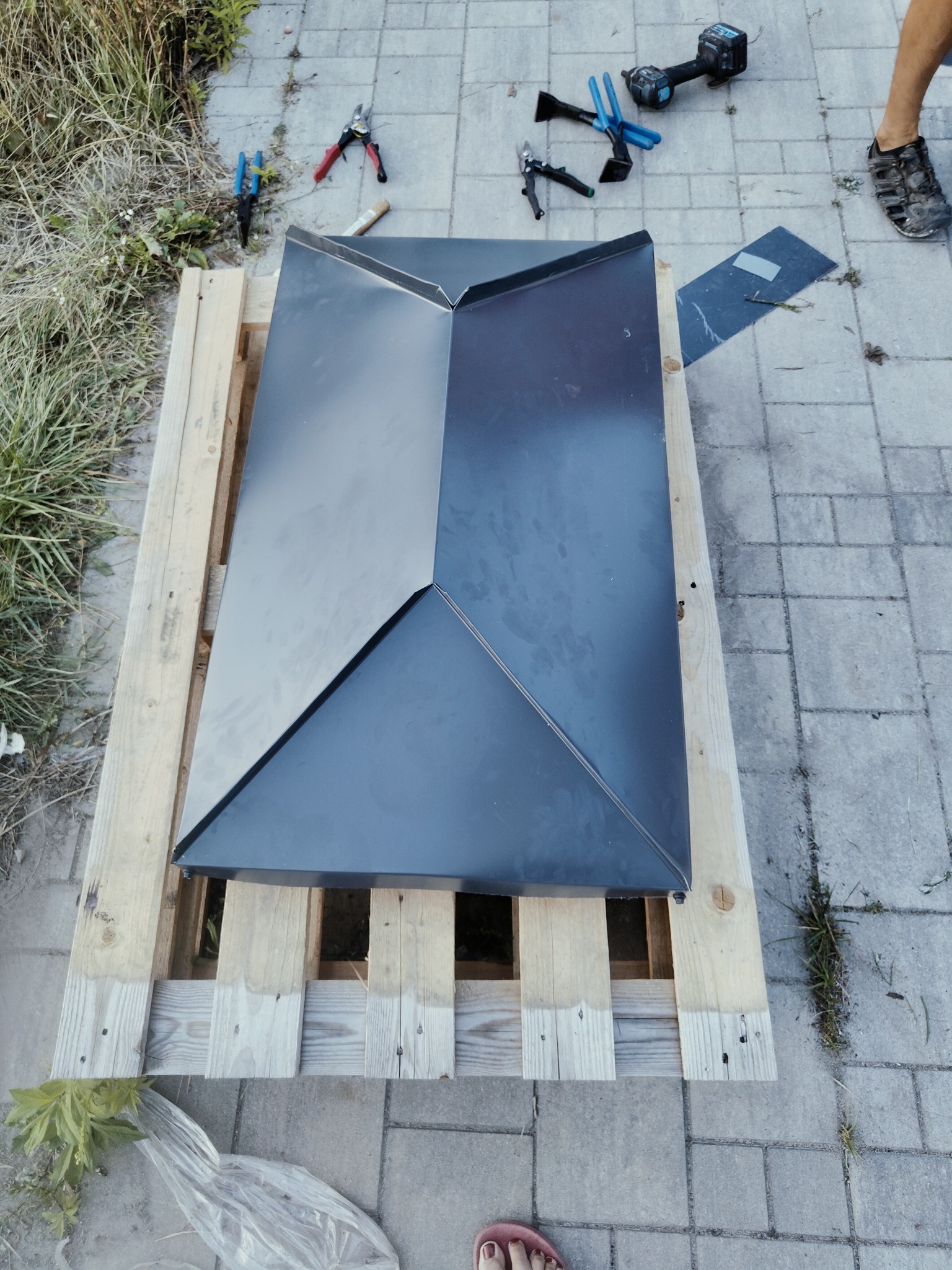 Gotowy element obróbki blacharskiej w kolorze grafitowym, ułożony na drewnianej palecie, z narzędziami blacharskimi w tle, na kostce brukowej.