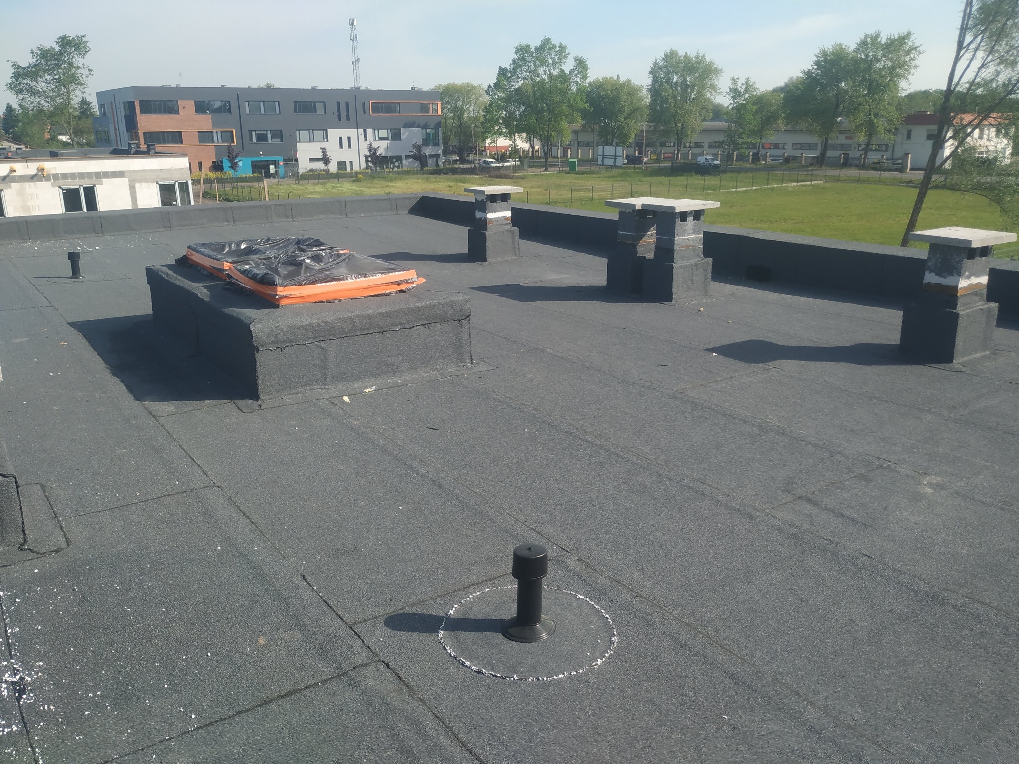 Pokrycie dachu papą termozgrzewalną na budynku z kominami, zabezpieczone okno dachowe folią i taśmą. Widoczny budynek w tle.