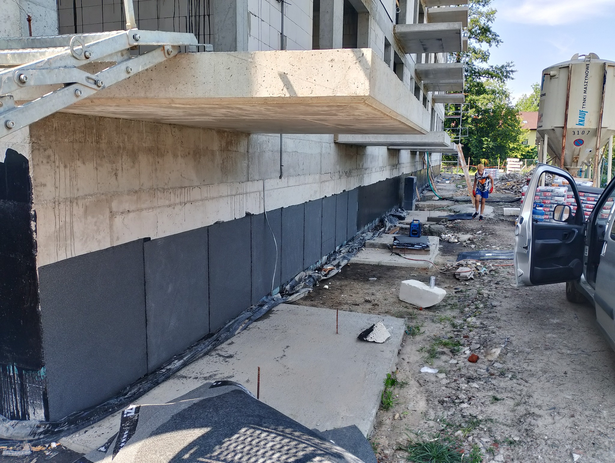 Czarna izolacja pionowa fundamentów budynku wielorodzinnego w trakcie realizacji, widoczna osoba i betoniarka Knauf w tle.