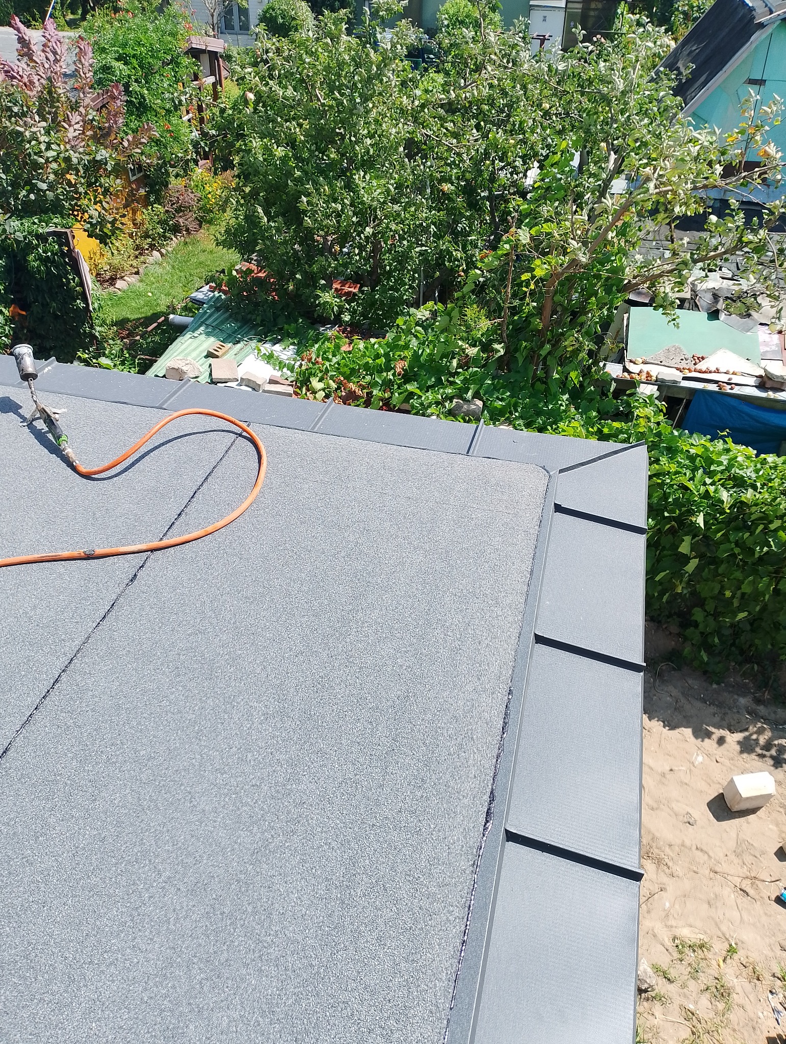 Nowe pokrycie dachu z papy termozgrzewalnej w kolorze szarym, widoczny palnik dekarski z przewodem oraz fragment ogrodu w tle.