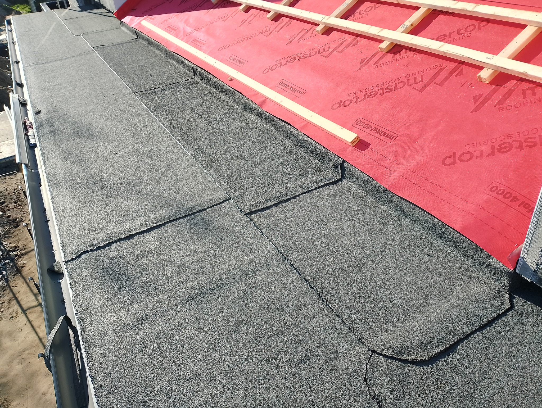 Fragment dachu płaskiego pokrytego papą, z widocznymi łączeniami i fragmentem membrany dachowej z drewnianą konstrukcją pod przyszłe warstwy dachu.