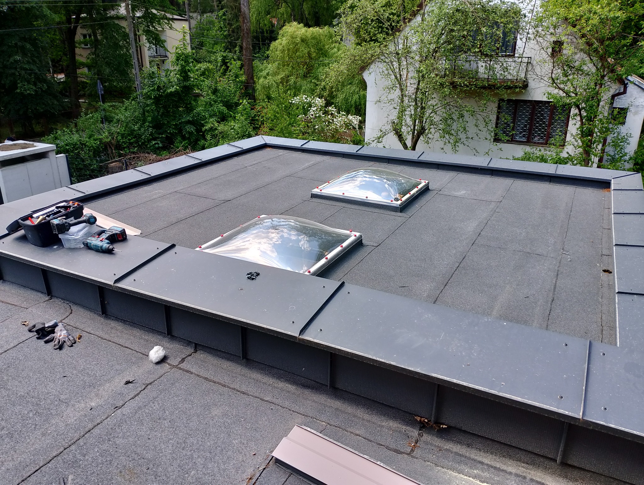 Nowy, płaski dach z papy z dwoma świetlikami kopułowymi; widoczny zestaw narzędzi dekarskich, rękawice robocze i obróbki blacharskie.