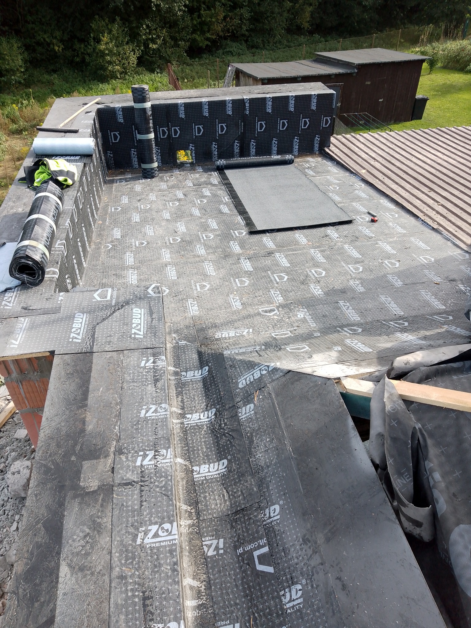 Płaski dach pokryty papą termozgrzewalną z widocznymi rolkami materiału, fragmentami muru i narzędziami, w tle budynki gospodarcze i zieleń.