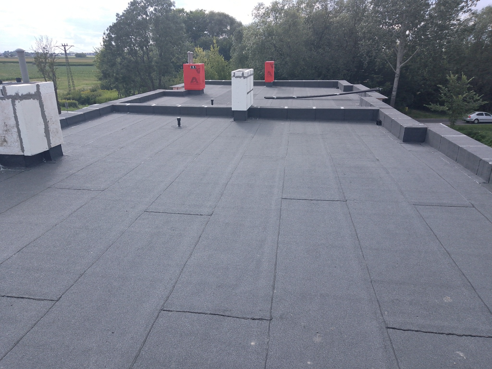 Widok z góry na płaski dach pokryty szarą papą termozgrzewalną z widocznymi kominami zabezpieczonymi czerwoną folią i obróbkami blacharskimi, w tle zieleń drzew i zabudowania.