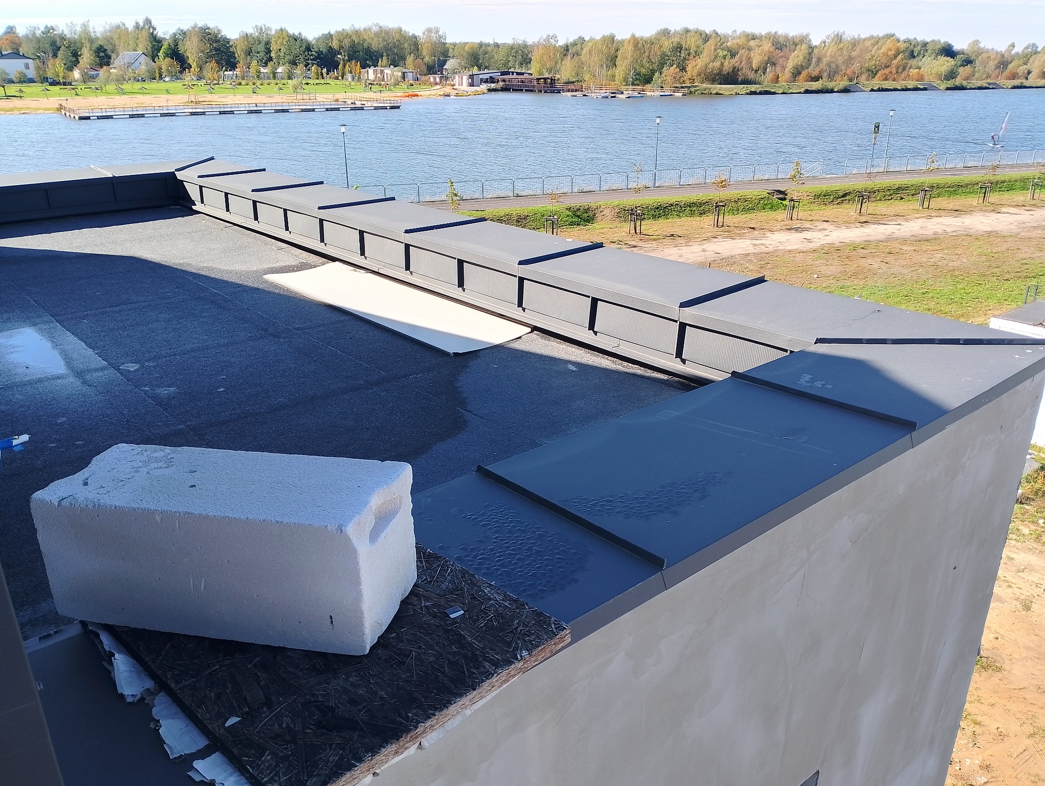 Widok z góry na fragment płaskiego dachu z obróbkami blacharskimi w kolorze grafitowym, przy krawędzi leży bloczek z betonu komórkowego, w tle jezioro i zabudowania.