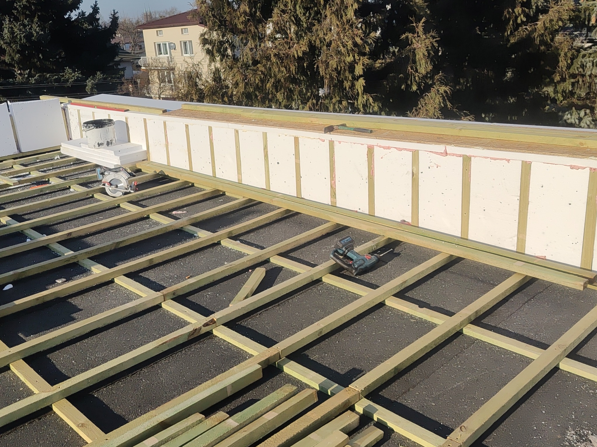 Konstrukcja drewniana na dachu z widoczną izolacją termiczną i narzędziem do cięcia, przygotowana pod dalsze prace dekarskie, w tle budynek mieszkalny i drzewa.