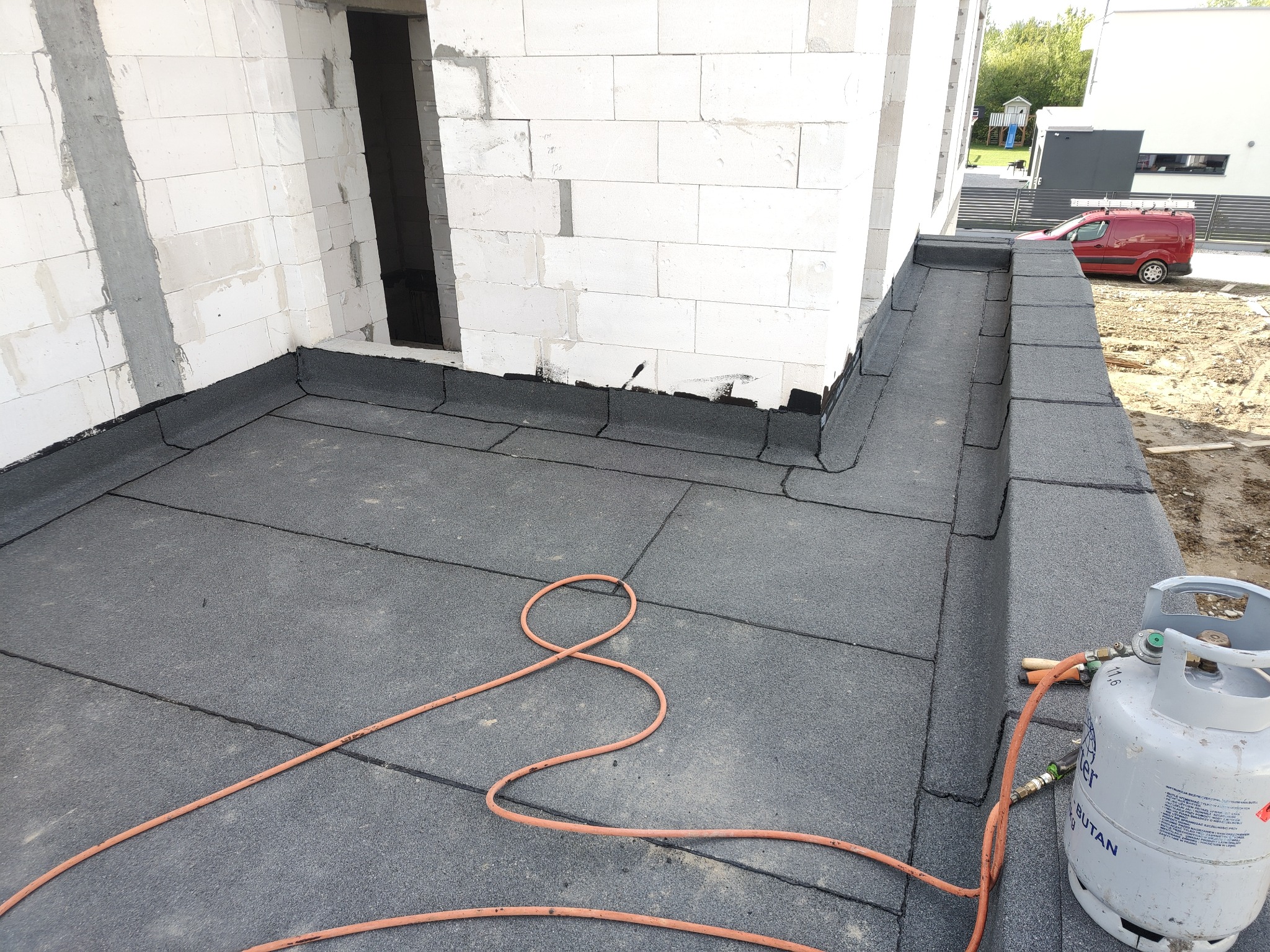 Szczelna izolacja dachu płaskiego z papy termozgrzewalnej, widoczna butla gazowa i palnik dekarski, ściany budynku z betonu komórkowego.