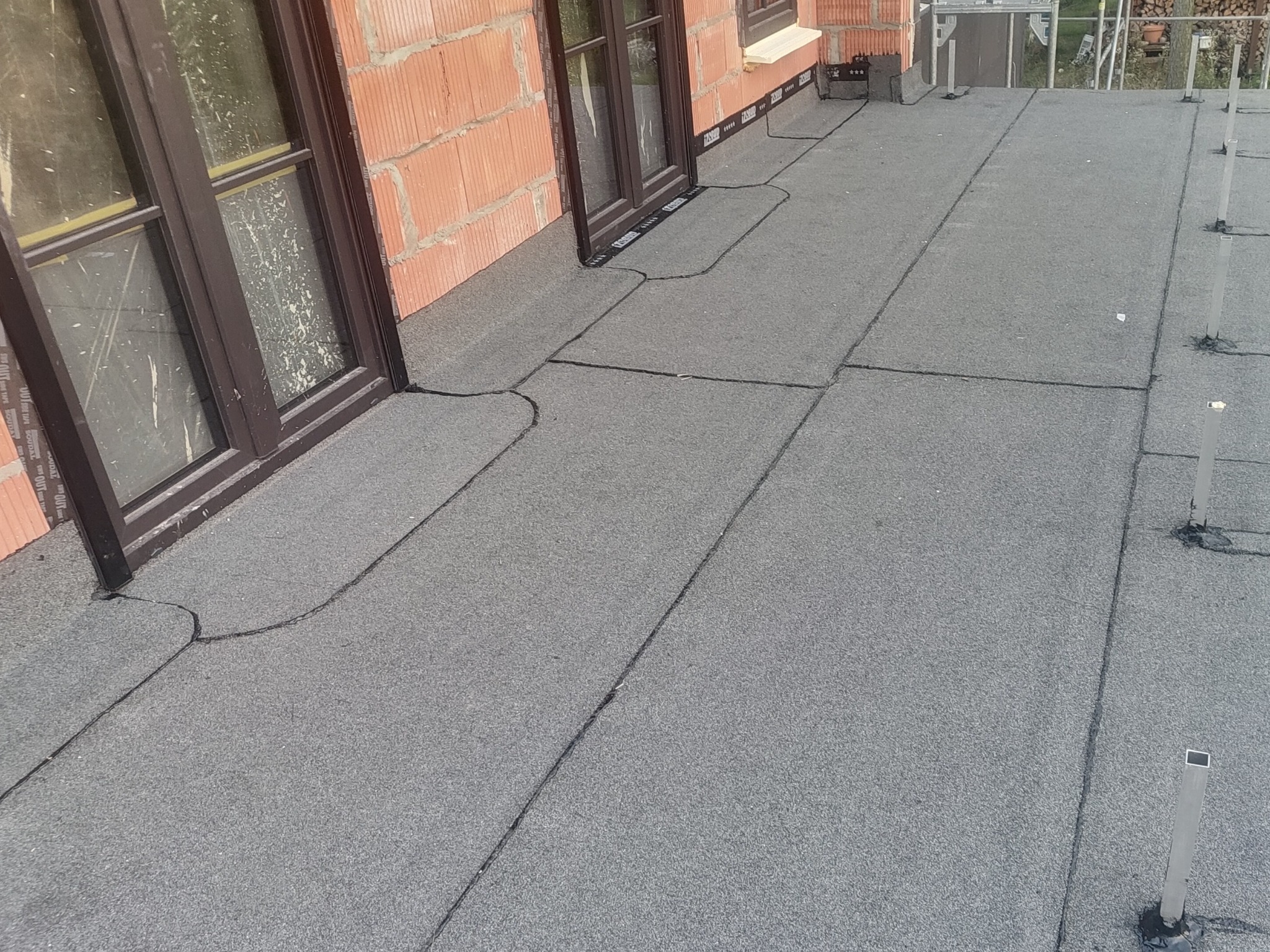 Szczegółowe ujęcie hydroizolacji na płaskim dachu z oknami, widoczne łączenia papy i elementy mocujące, ceglana ściana budynku w tle.