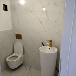 Invest Bud Karol Baranowski - Biała umywalka kolumnowa z złotą baterią, wisząca toaleta z deską wolnoopadającą, marmurowe płytki na ścianach i podłodze, włącznik światła w kolorze mosiądzu, surowy otwór na gniazdko elektryczne.