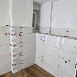 Invest Bud Karol Baranowski - Układanie jasnych, podłużnych płytek na ścianach łazienki z użyciem czerwonych krzyżyków i białej taśmy, widoczne otwory pod instalacje, drewnopodobna podłoga, surowy sufit z widocznymi śladami...