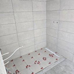 Invest Bud Karol Baranowski - Jasnoszare płytki ceramiczne na ścianach i podłodze, z systemem poziomowania płytek (czerwone klipsy i białe kliny) na podłodze, widoczne przewody instalacji elektrycznej w rogu pomieszczenia.