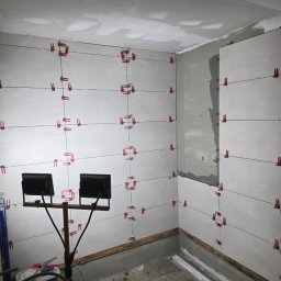 Invest Bud Karol Baranowski - Ściany łazienki w trakcie układania płytek, widoczne krzyżyki dystansowe, oświetlenie robocze i przygotowane podłoże.