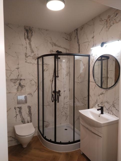 Nowoczesna łazienka z kabiną prysznicową w czarnej ramie, umywalką z szafką i okrągłym lustrem na ścianie wyłożonej płytkami imitującymi marmur.