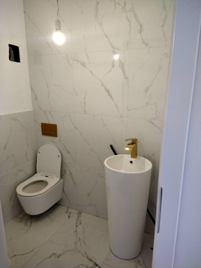 Biała umywalka kolumnowa z złotą baterią, wisząca toaleta z deską wolnoopadającą, marmurowe płytki na ścianach i podłodze, włącznik światła w kolorze mosiądzu, surowy otwór na gniazdko elektryczne.