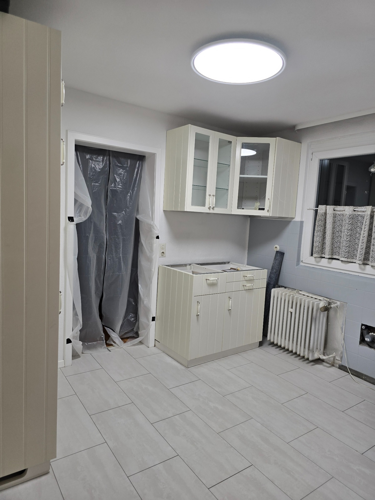 Kuchnia w trakcie prac: zabezpieczone folią drzwi, częściowo zdemontowane meble, widoczne płytki podłogowe i grzejnik.
