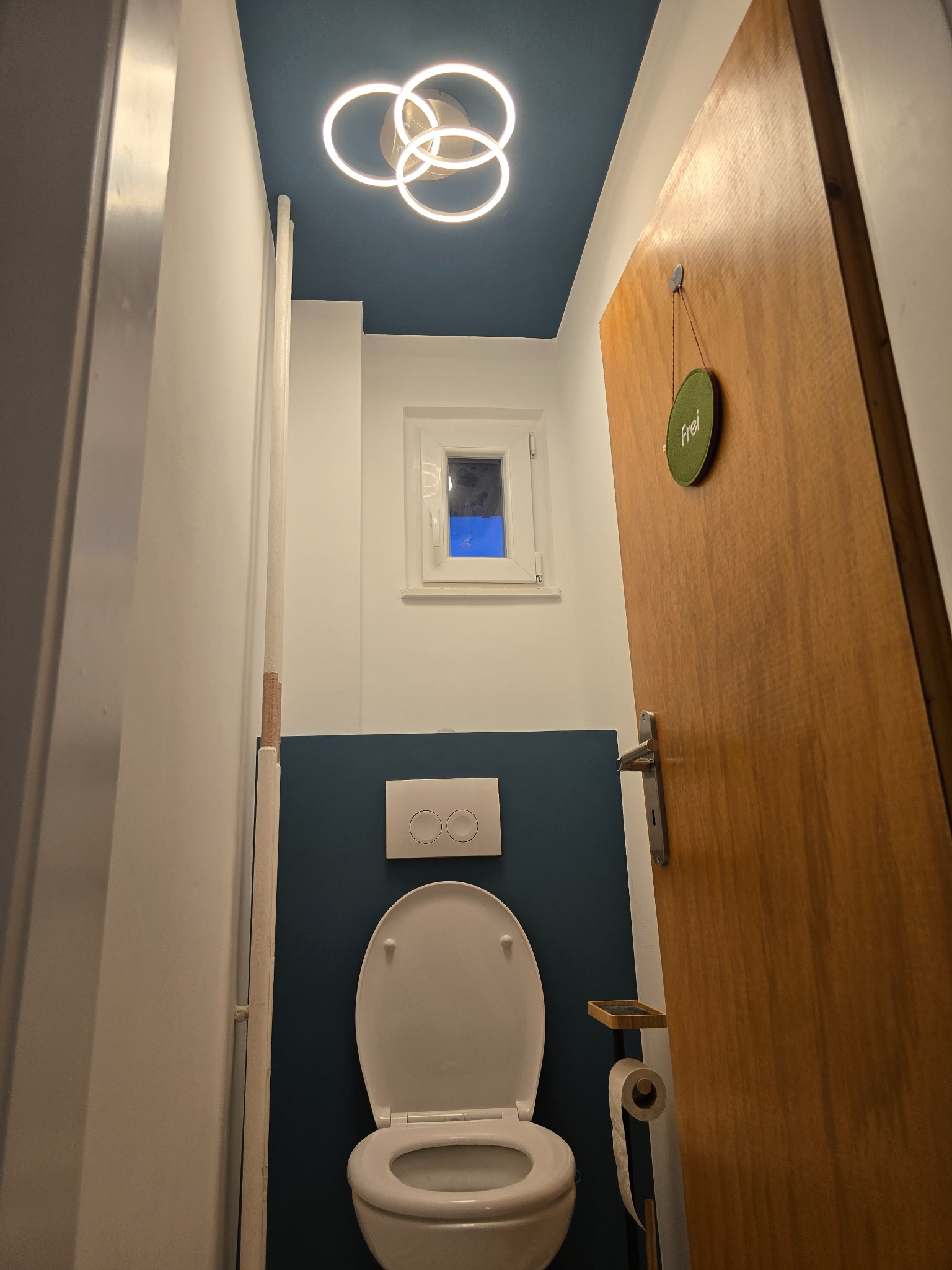 Wnętrze małej łazienki z otwartą drewnianą toaletą, niebieską ścianą, nowoczesną lampą sufitową w kształcie połączonych okręgów i małym oknem z widokiem na nocne niebo.