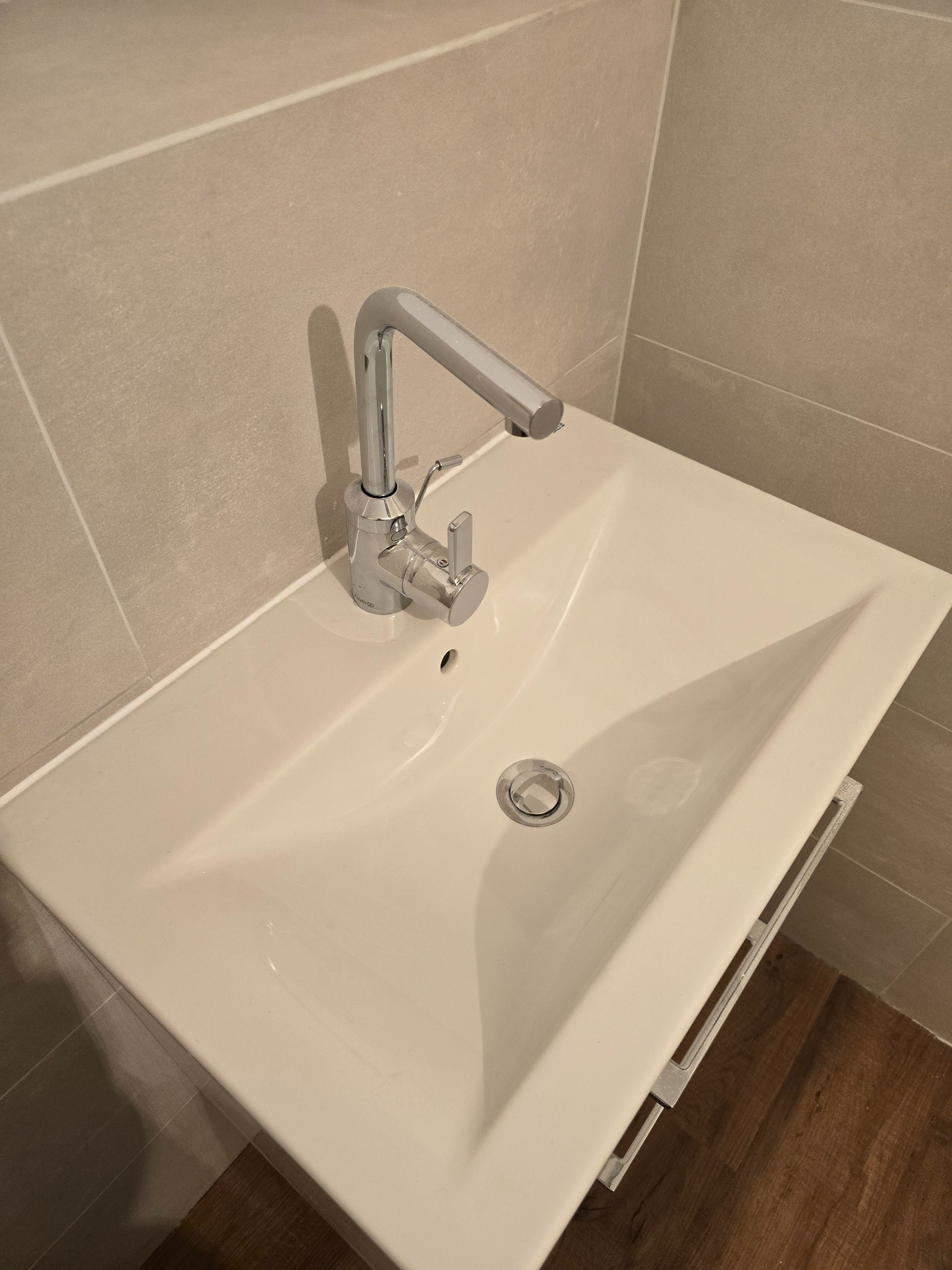 Biała umywalka z chromowaną baterią w nowoczesnej łazience z beżowymi płytkami i drewnopodobną podłogą, widok z góry.