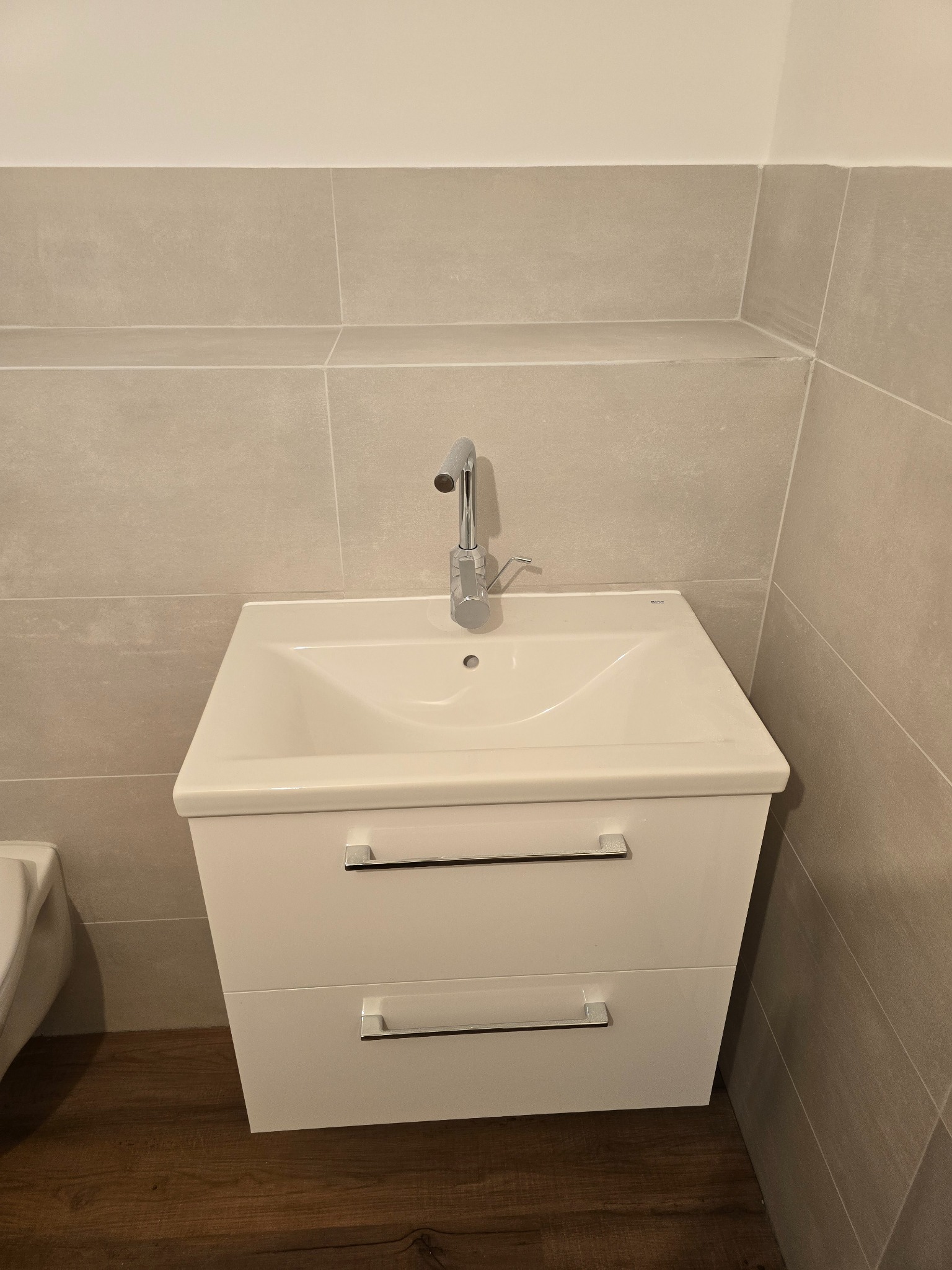 Nowoczesna biała umywalka z szafką i chromowaną baterią, zamontowana na tle płytek w odcieniach szarości i podłogi imitującej drewno.