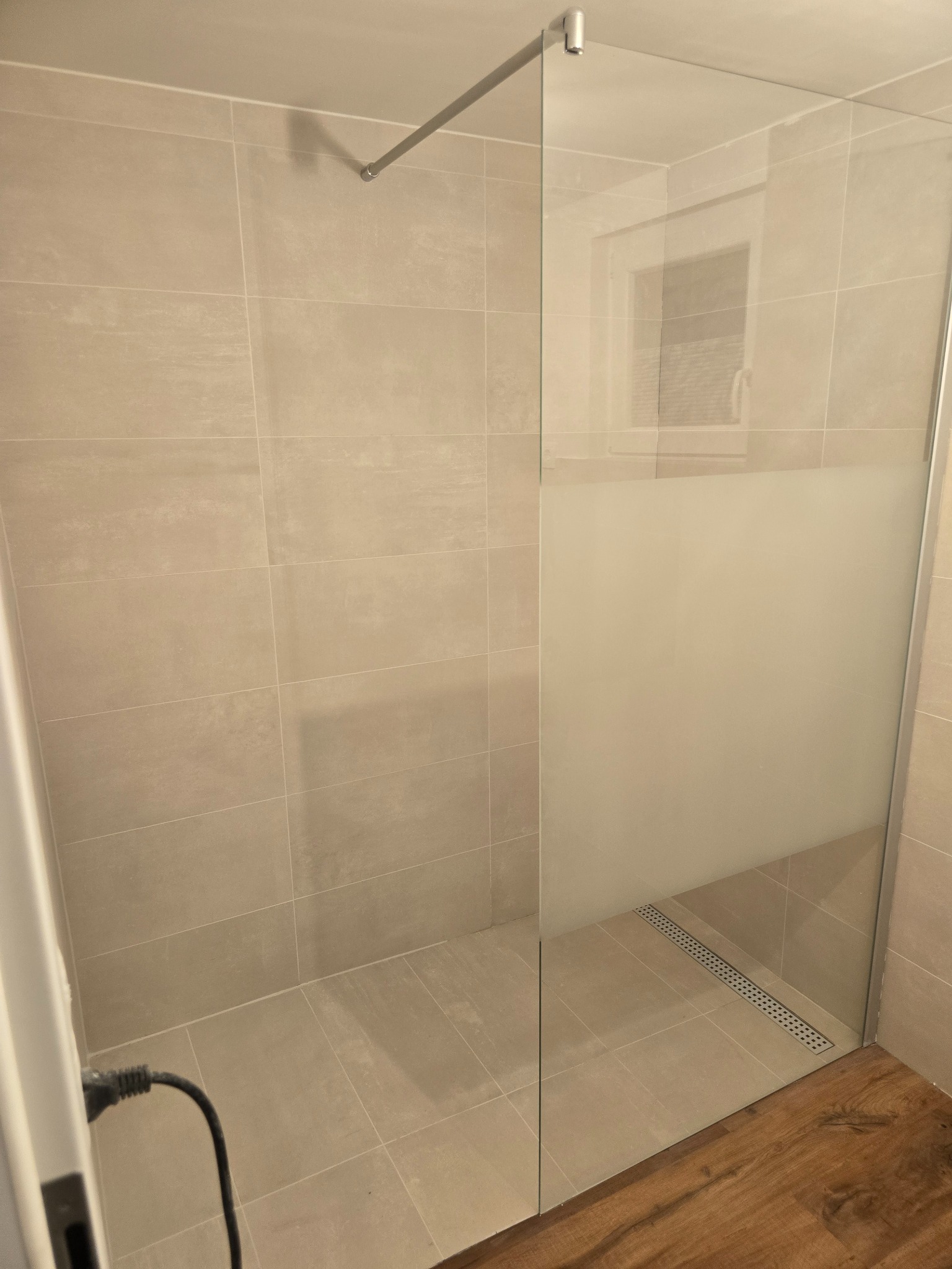 Beżowa łazienka z kabiną prysznicową typu walk-in, częściowo matową szybą i odpływem liniowym w podłodze wyłożonej płytkami; drewnopodobna podłoga na zewnątrz kabiny.