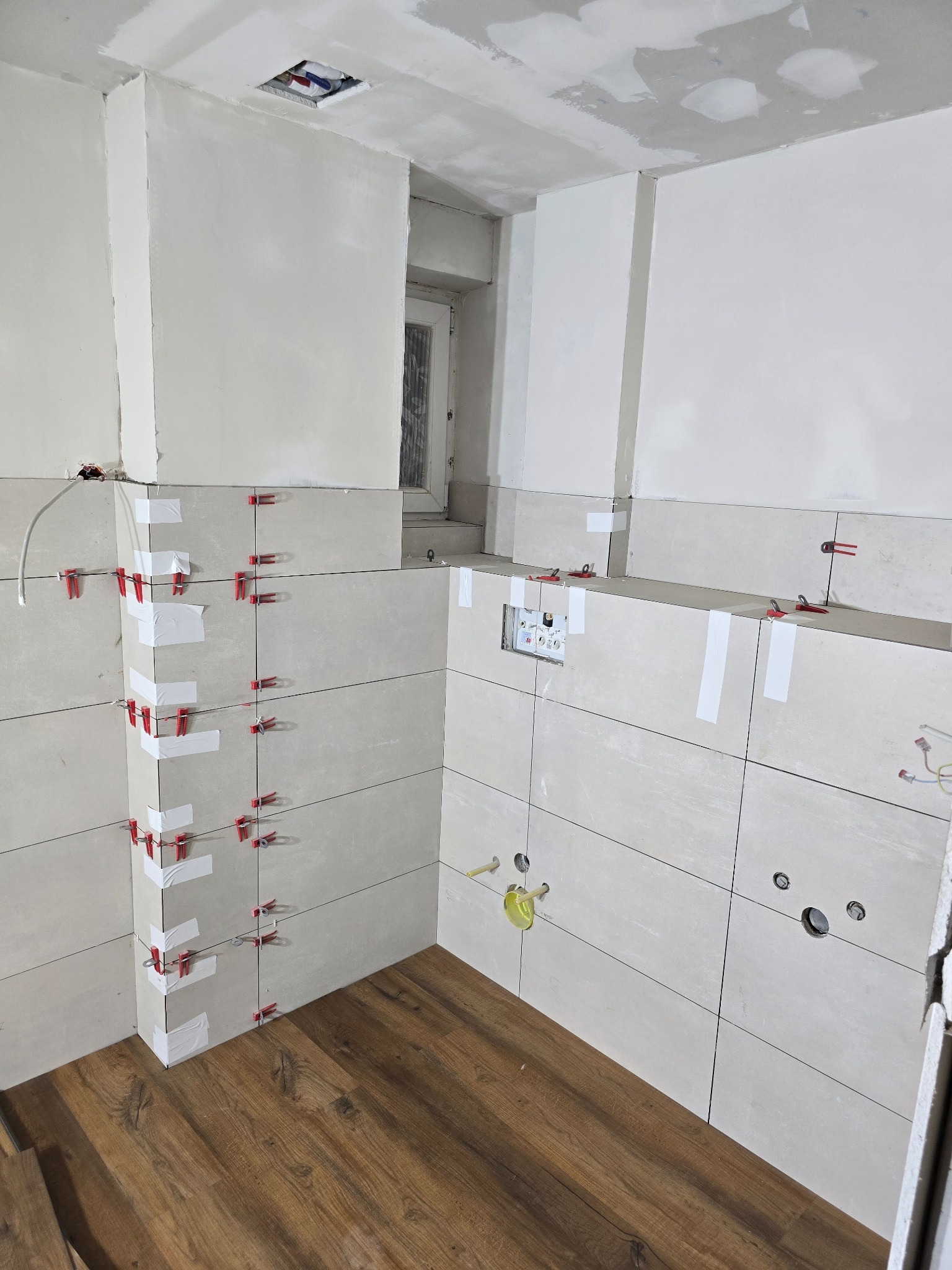 Układanie jasnych, podłużnych płytek na ścianach łazienki z użyciem czerwonych krzyżyków i białej taśmy, widoczne otwory pod instalacje, drewnopodobna podłoga, surowy sufit z widocznymi śladami...