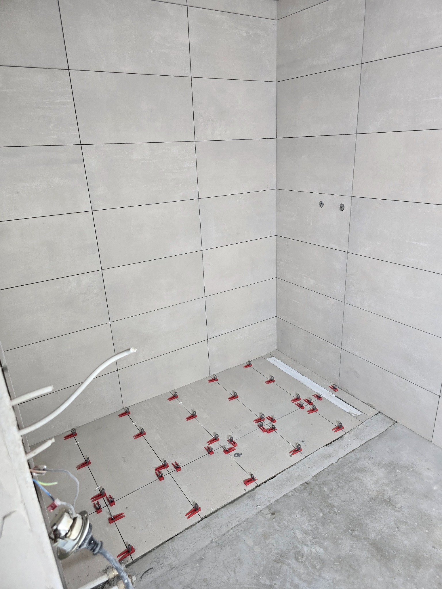 Jasnoszare płytki ceramiczne na ścianach i podłodze, z systemem poziomowania płytek (czerwone klipsy i białe kliny) na podłodze, widoczne przewody instalacji elektrycznej w rogu pomieszczenia.
