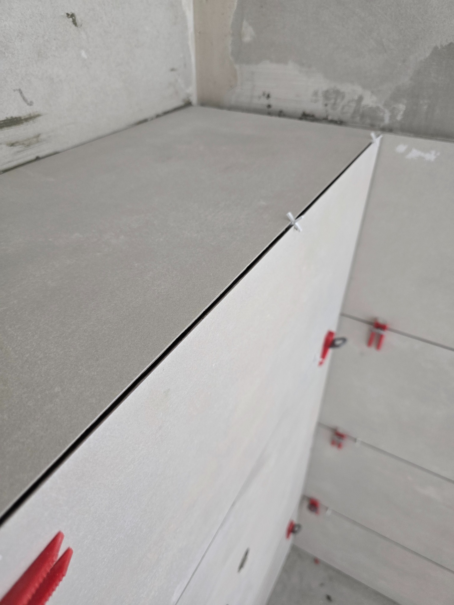 Świeżo ułożone, szare płytki ścienne w rogu pomieszczenia, z krzyżykami dystansowymi w kolorze białym i czerwonym, na tle surowego, nieotynkowanego sufitu.