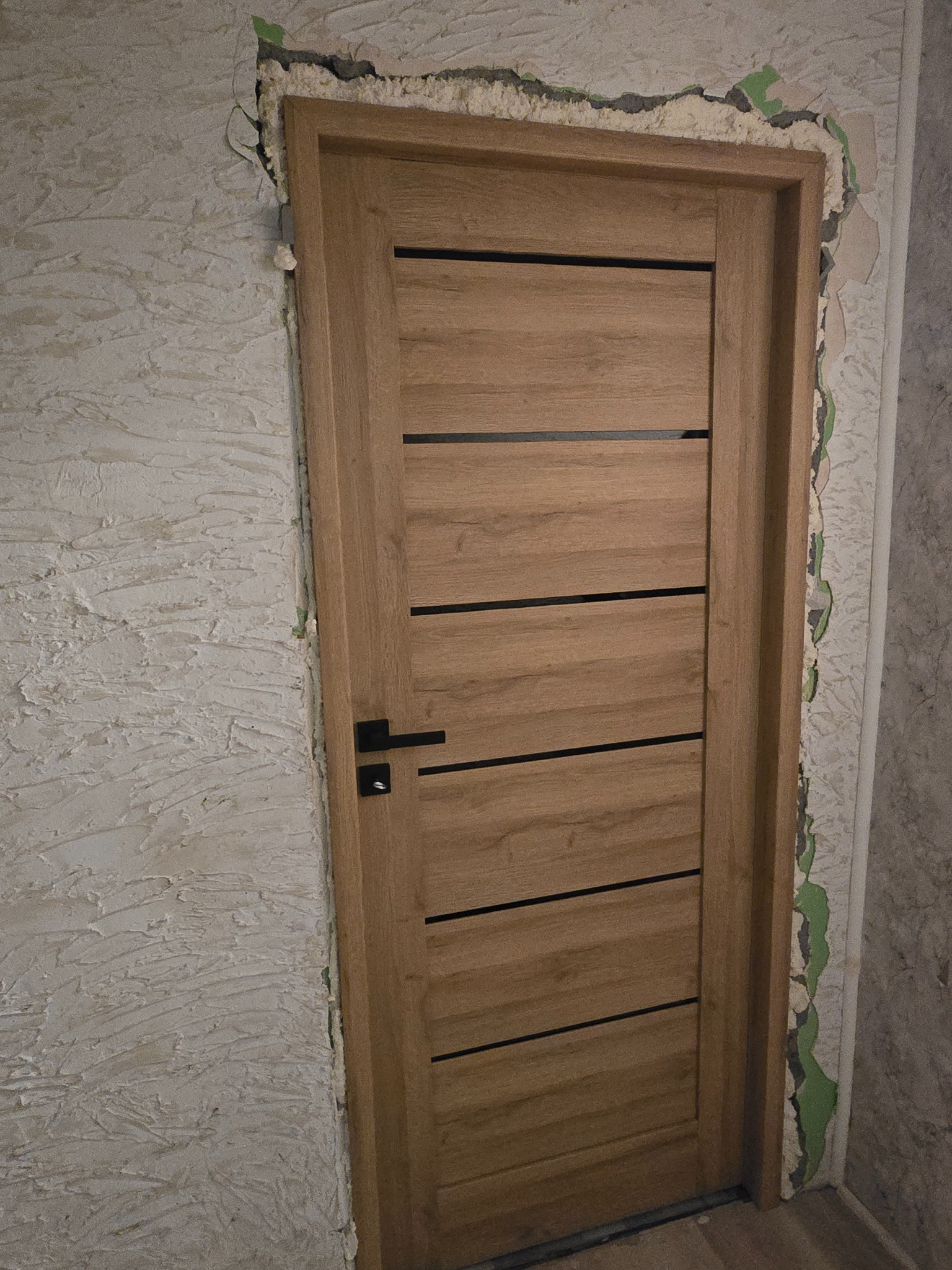 Dębowa wewnętrzna drzwi z czarnymi poziomymi akcentami, czarną klamką i ościeżnicą, w trakcie montażu, widoczne ślady pianki montażowej i niedokończone wykończenie ściany.