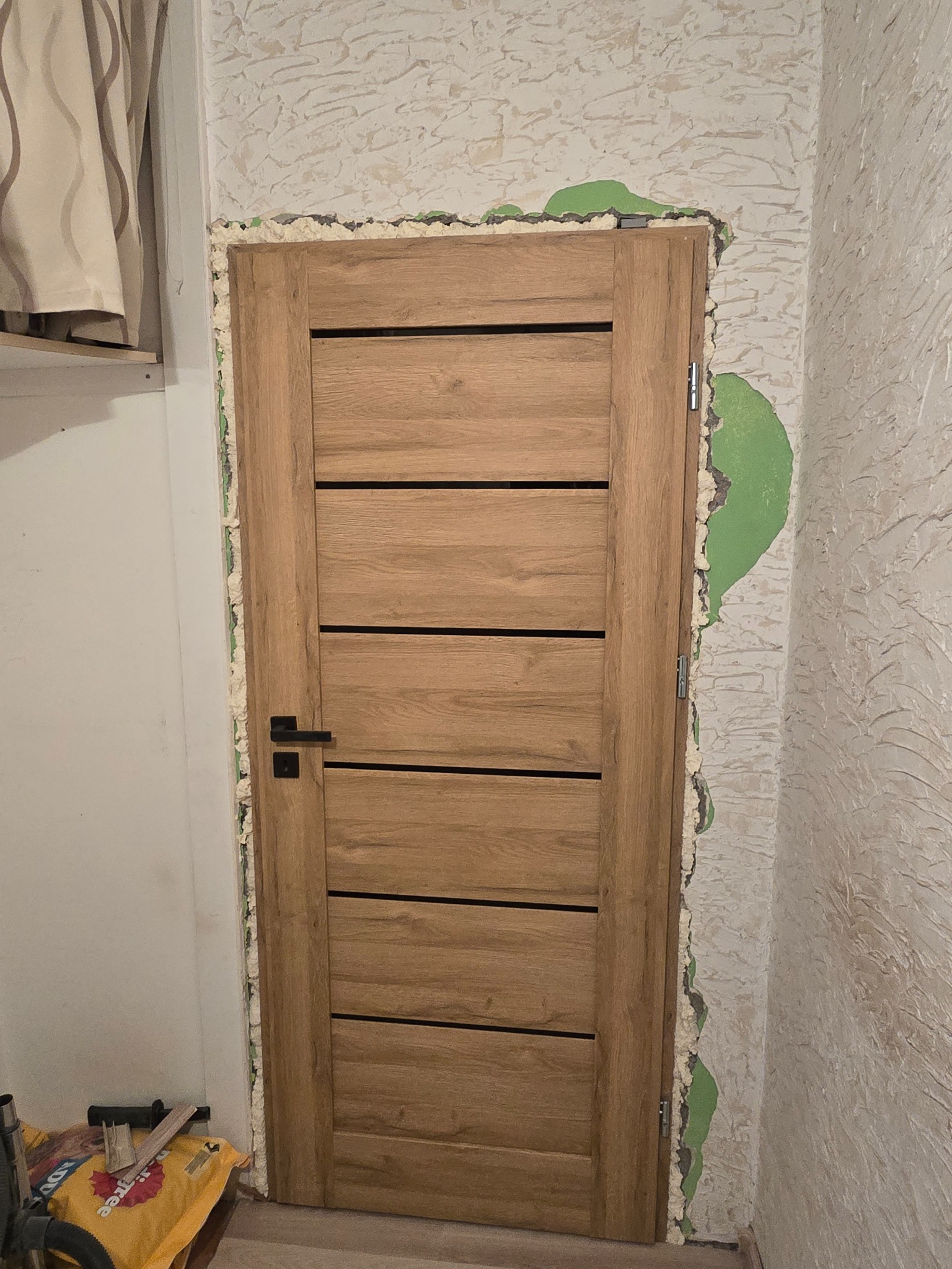 Dębowa, nowoczesna drzwi wewnętrzne z czarną klamką, osadzona w ościeżnicy z widoczną pianką montażową i fragmentami zielonej farby na ścianie o teksturowanej powierzchni.