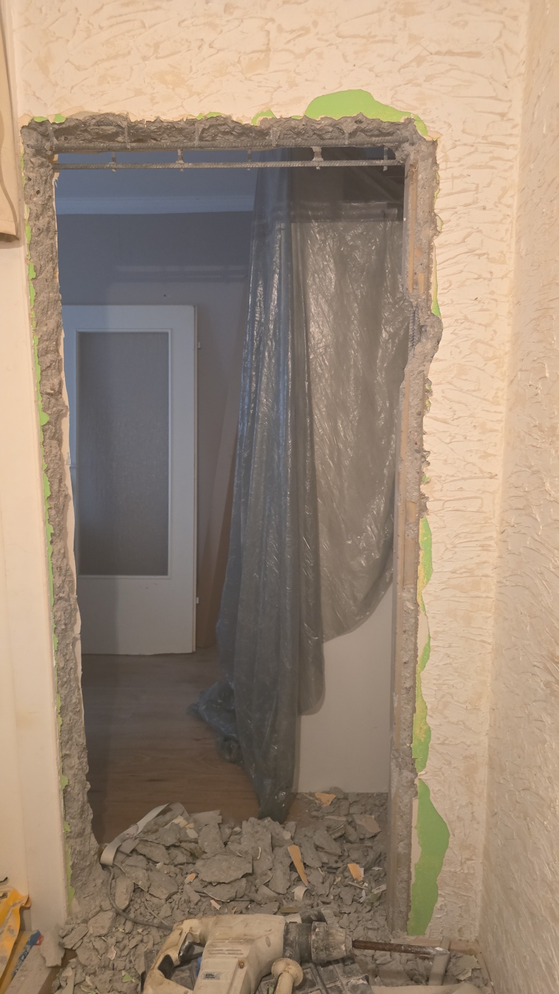 Otwór w ścianie przygotowany do montażu ościeżnicy, z widocznym zbrojeniem i resztkami gruzu oraz narzędziem na podłodze, zabezpieczony folią malarską.