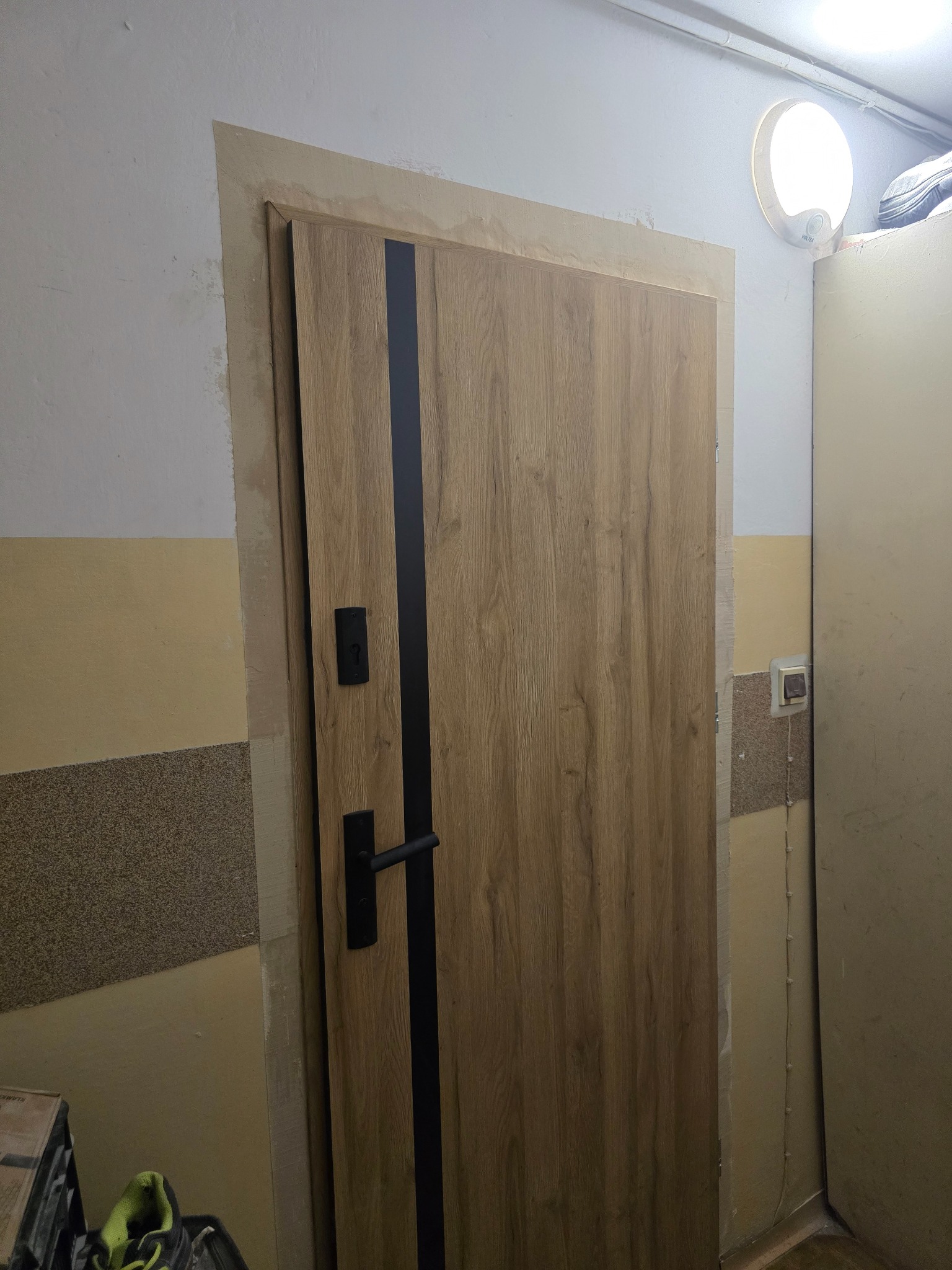 Drzwi wewnętrzne z okleiną drewnopodobną i czarnym paskiem, zamontowane w ościeżnicy, widoczne ślady prac wykończeniowych na ścianie i ościeżnicy.
