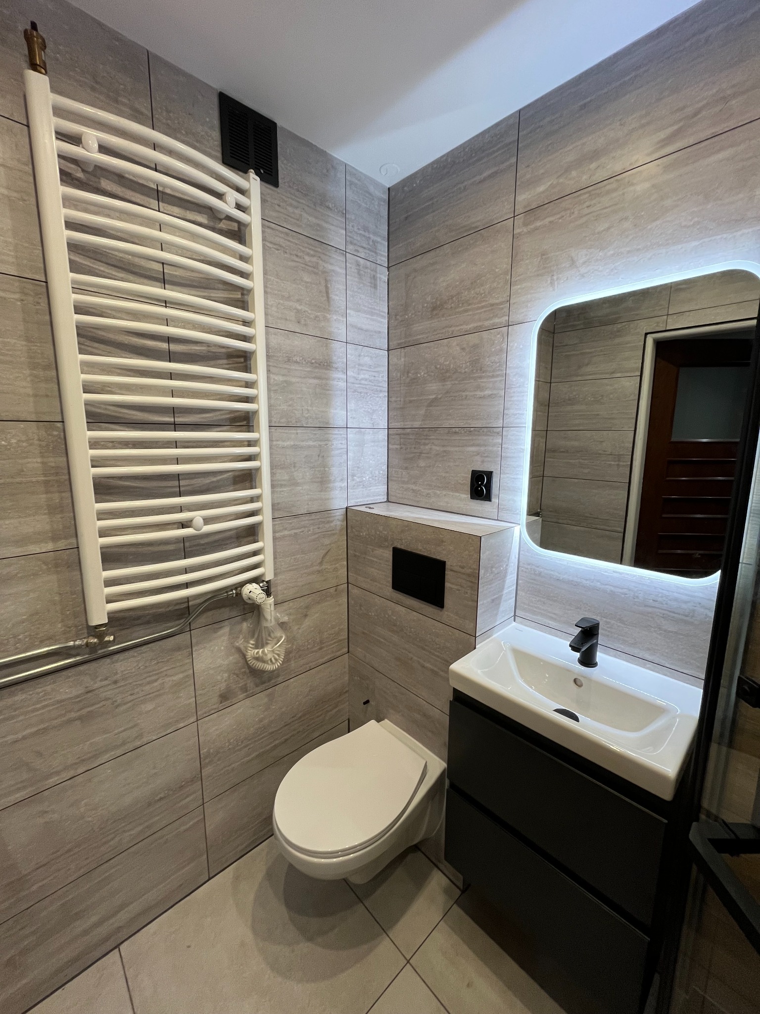 Nowoczesna łazienka z szarymi płytkami imitującymi drewno, podświetlanym lustrem, białą umywalką z czarną armaturą i grzejnikiem drabinkowym.