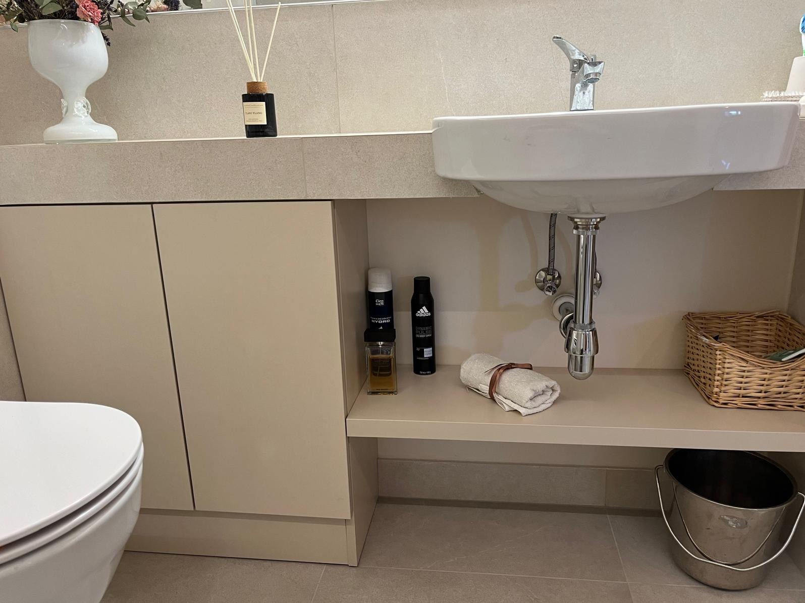 Nowoczesna łazienka z beżowymi szafkami pod umywalką, białą ceramiką i dekoracyjnymi akcentami, takimi jak zapachowe patyczki i wiklinowy koszyk.