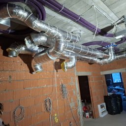 InstalMax Jarosław Gunarski - Rozbudowany system kanałów wentylacyjnych w folii aluminiowej i fioletowych rur karbowanych zamontowany pod surowym betonowym stropem w budynku w trakcie budowy, widoczne ceglane ściany i zwisające...