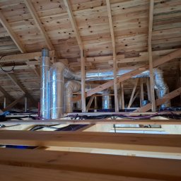 InstalMax Jarosław Gunarski - Widok instalacji wentylacyjnej z aluminiowymi rurami w surowym drewnianym poddaszu, widoczne kable i przewody.