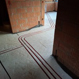 InstalMax Jarosław Gunarski - Czerwone rury instalacji grzewczej ułożone w bruzdach w podłodze z betonu komórkowego, widoczne ściany z pustaków ceramicznych z wyprowadzonymi przewodami elektrycznymi.