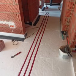 InstalMax Jarosław Gunarski - Czerwone rury układane równolegle na białym styropianie pod ogrzewanie podłogowe w trakcie montażu, widoczne ceglane ściany w surowym stanie, narzędzia i wiadro z zaprawą w tle.