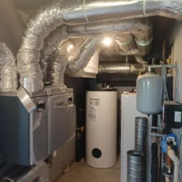 Instalacja rekuperacji z centralą wentylacyjną w kotłowni, widoczne liczne elastyczne przewody wentylacyjne, zbiornik buforowy i elementy instalacji hydraulicznej przy ścianie.