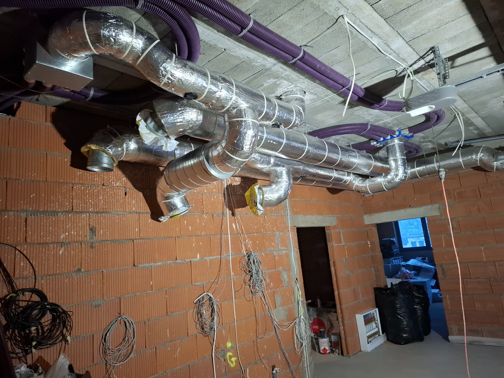 Rozbudowany system kanałów wentylacyjnych w folii aluminiowej i fioletowych rur karbowanych zamontowany pod surowym betonowym stropem w budynku w trakcie budowy, widoczne ceglane ściany i zwisające...