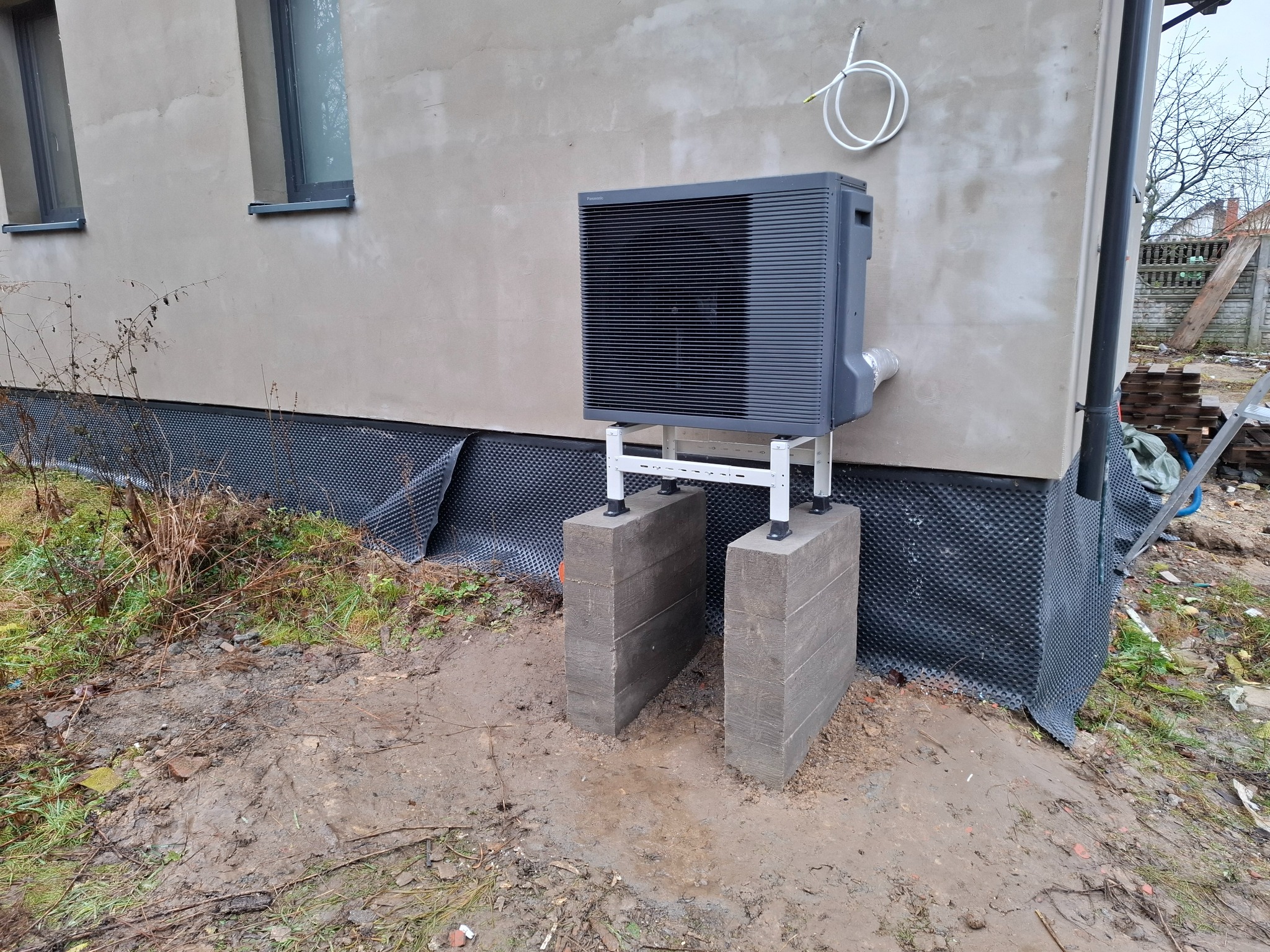 Zewnętrzna jednostka szarej pompy ciepła Panasonic zamontowana na betonowych blokach przy ścianie budynku z widoczną folią kubełkową i błotnistym terenem.