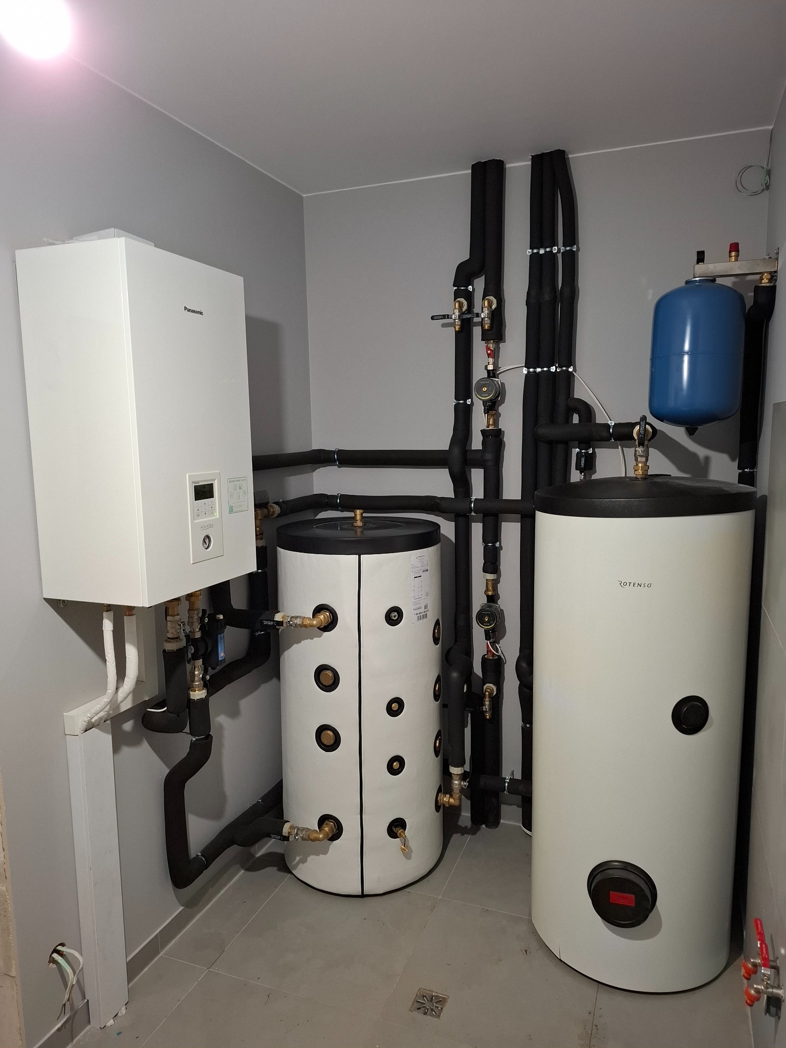 Nowoczesna kotłownia z białym piecem gazowym Panasonic Aquarea, dwoma białymi zbiornikami Rotenso i czarną izolacją na rurach w szarym pomieszczeniu.