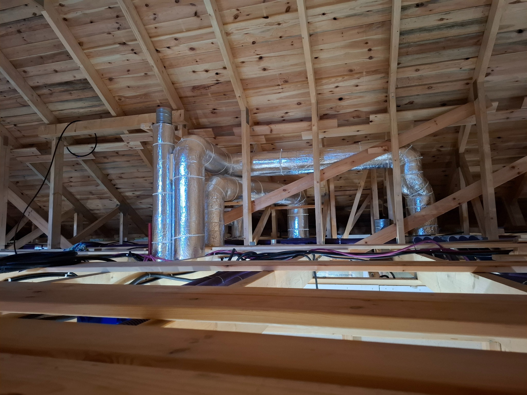 Widok instalacji wentylacyjnej z aluminiowymi rurami w surowym drewnianym poddaszu, widoczne kable i przewody.