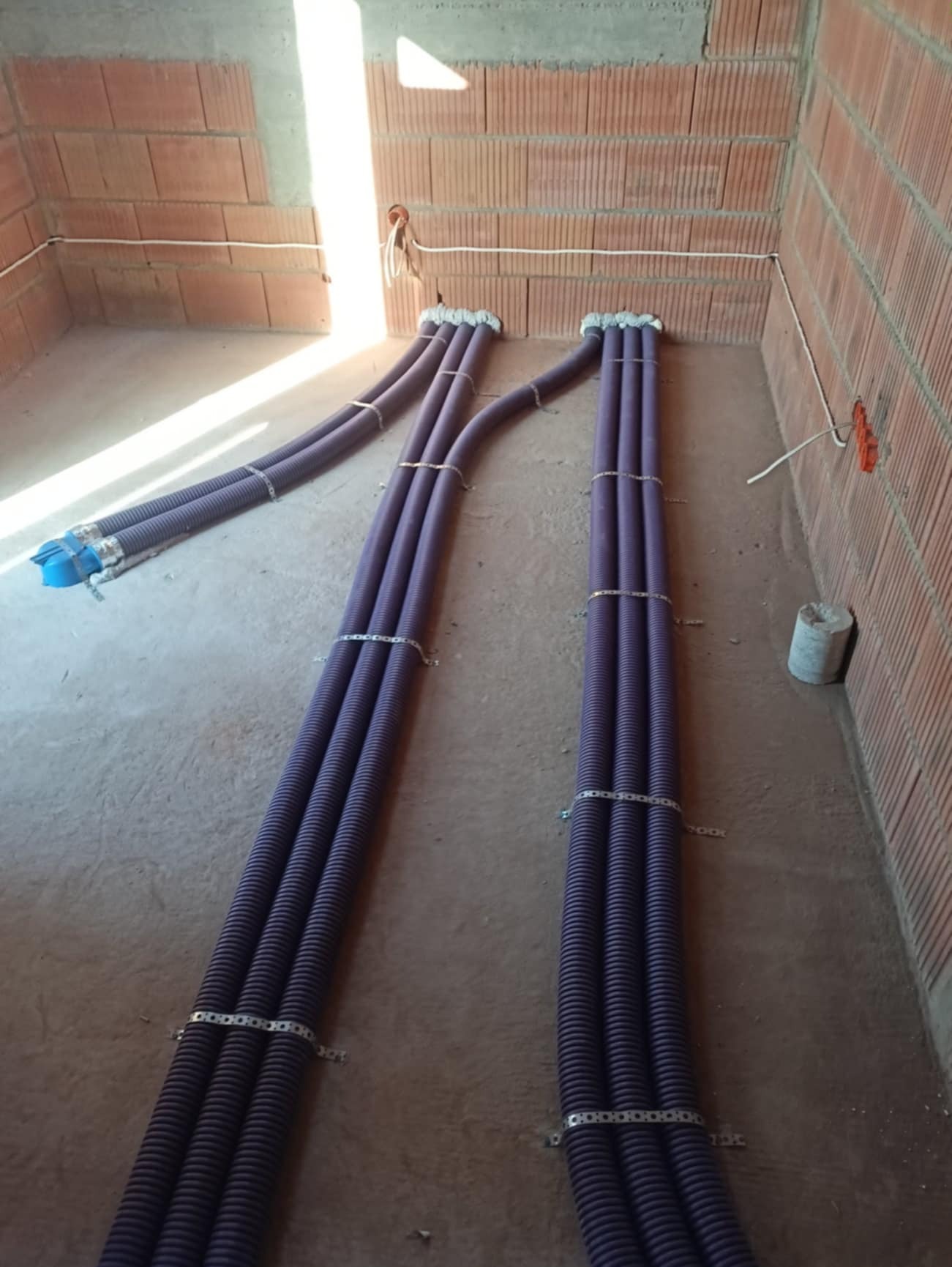 Fioletowe rury instalacyjne, zebrane w wiązki i przymocowane metalowymi obejmami, prowadzone w surowym wnętrzu z ceglanymi ścianami i betonową posadzką, widoczne przewody elektryczne.