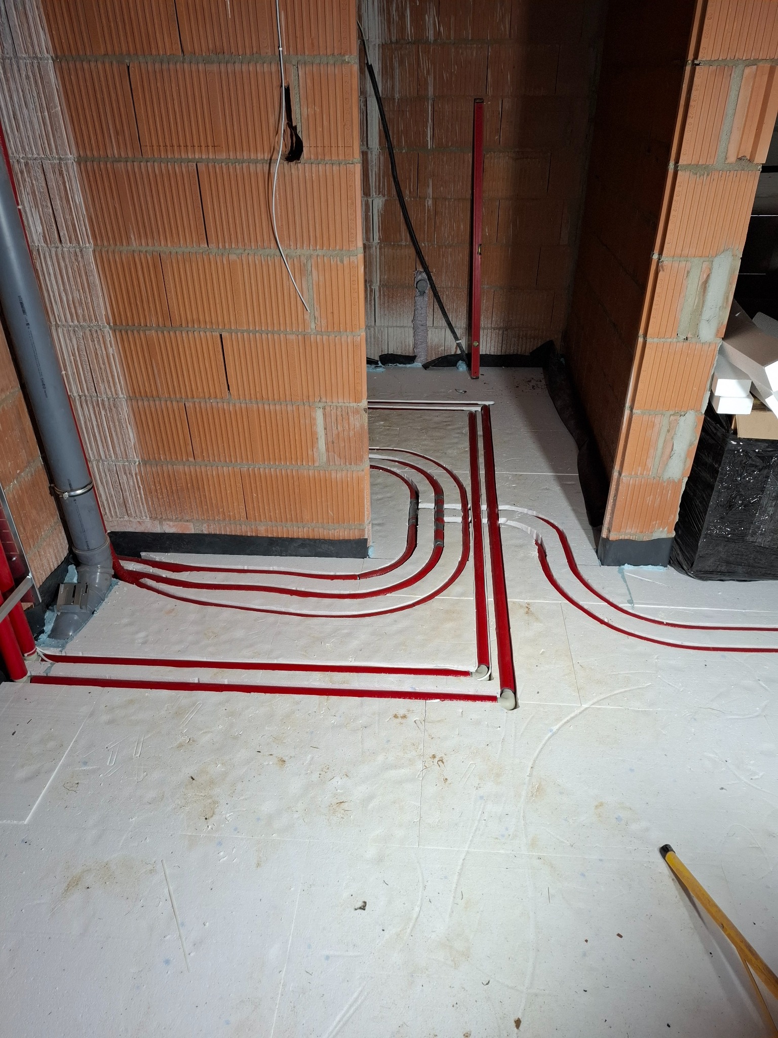 Czerwone rury grzewcze ułożone na białym styropianie podłogowym, widoczne ceglane ściany w surowym stanie, poziomica oparta o ścianę.